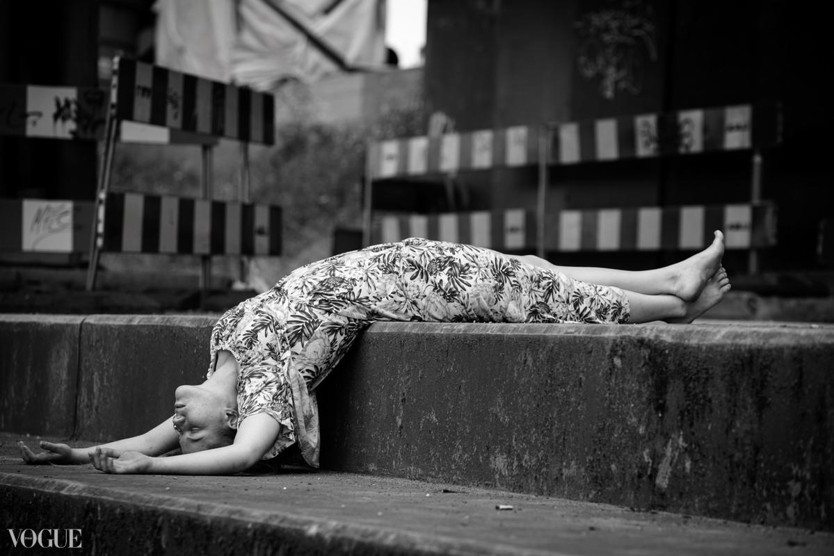 Czarno-białe zdjęcie kobiety w kwiecistej sukience, leżącej z głową w dół na betonowej konstrukcji w miejskiej scenerii z barierami w tle, stylizowane na sesję modową z logo 'VOGUE' w lewym dolnym...