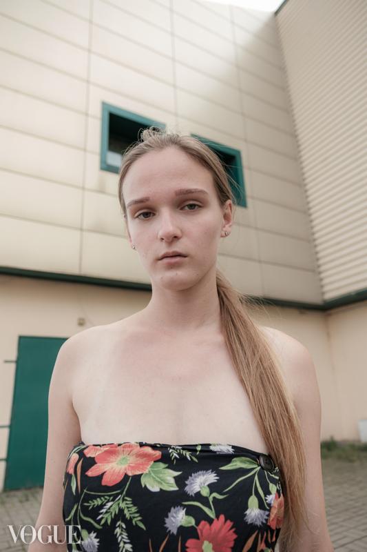 Portret młodej kobiety z długimi blond włosami, ubrana w kwiecistą sukienkę bez ramiączek, na tle budynku z małymi oknami.