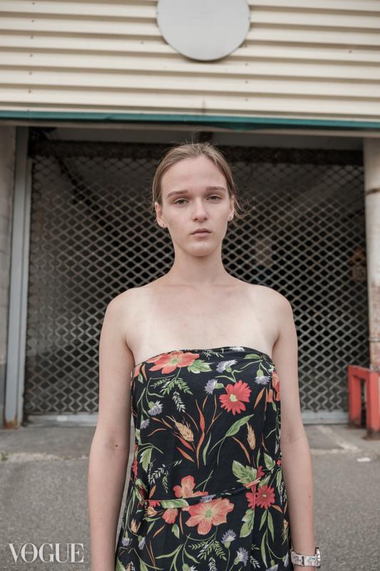 Portret młodej kobiety w sukience w kwiaty na tle zamkniętej, metalowej bramy z widocznym logo Vogue.