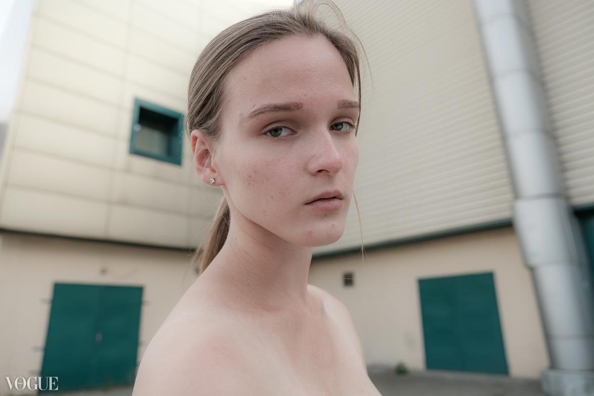 Portret kobiety z włosami spiętymi w kucyk, na tle budynku z zielonymi drzwiami i srebrną rurą wentylacyjną.