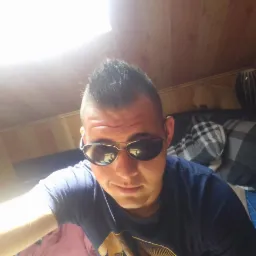 Mężczyzna w okularach przeciwsłonecznych, w pokoju z drewnianą boazerią i oknem dachowym, leżący na ł&oacute;żku z wzorzystą pościelą i poduszkami.