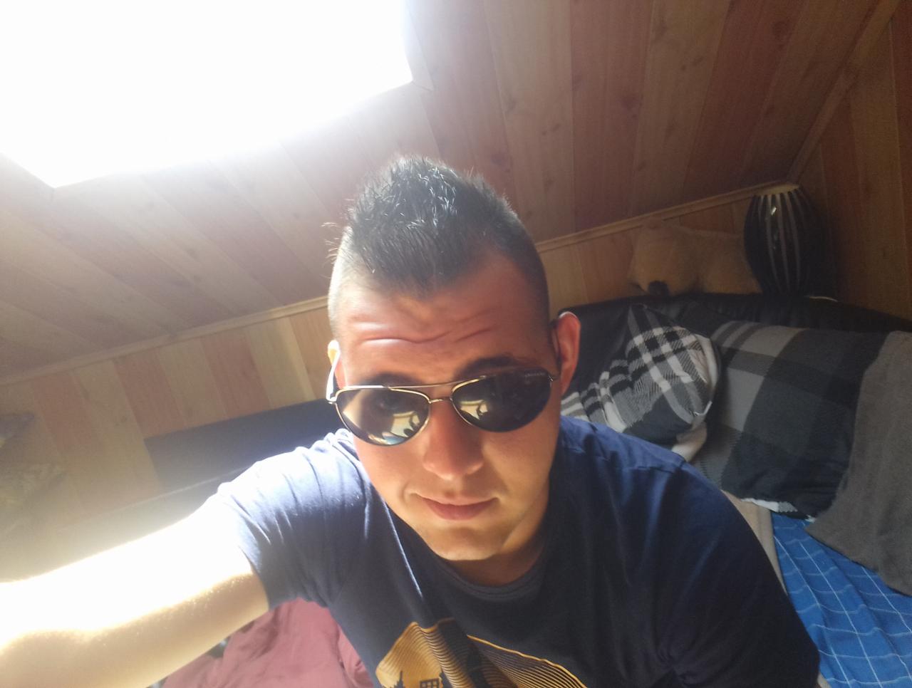 Mężczyzna w okularach przeciwsłonecznych, w pokoju z drewnianą boazerią i oknem dachowym, leżący na łóżku z wzorzystą pościelą i poduszkami.