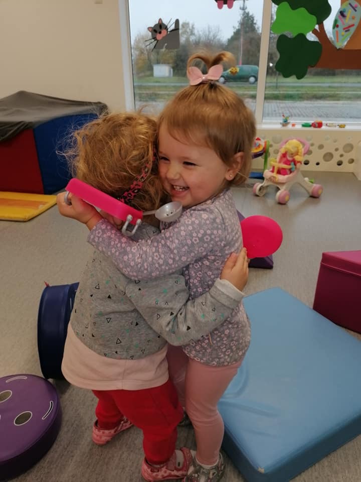 Dwie uśmiechnięte dziewczynki w ubraniach w pastelowych kolorach obejmują się w jasnym pokoju zabaw z zabawkami i naklejkami na oknie.