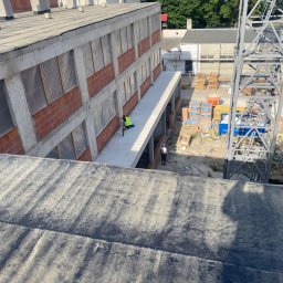 Budowlani - Wysoki kąt: Murarz w kamizelce, w trakcie prac murarskich na betonowym balkonie budynku w stanie surowym, z widoczną konstrukcją żurawia budowlanego.