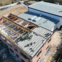 Budowlani - Budowa dwukondygnacyjnego budynku z cegły. Ekipa w kaskach układa prefabrykaty stropowe. Widok z góry na plac budowy i sąsiedni budynek z szarym dachem.