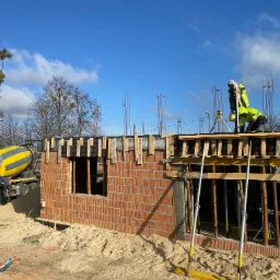 Budowa domu z betonu komórkowego: murarz w odblaskowej odzieży mierzy poziom, w tle pompa do betonu i błękitne niebo. Widoczne zbrojenia i drewniane szalunki.
