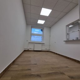 Budowlani - Wnętrze biura po remoncie: świeżo pomalowane ściany na biało, drewnopodobna podłoga, okno z kratą i grzejnikiem, sufit kasetonowy z oświetleniem.