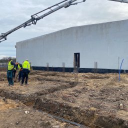 Budowlani - Budowa hali: fundamenty zbrojone stalą, pracownicy w odblaskowych kamizelkach, betonowa pompa w tle. Etap wylewania fundamentów pod budynek.