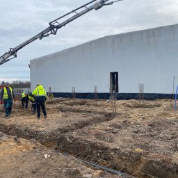 Budowlani - Budowa fundamentów budynku: ekipa w odblaskowych kamizelkach, betonowa pompa, wykopane rowy zbrojeniowe, w tle surowa ściana budynku. Prace ziemne w toku.