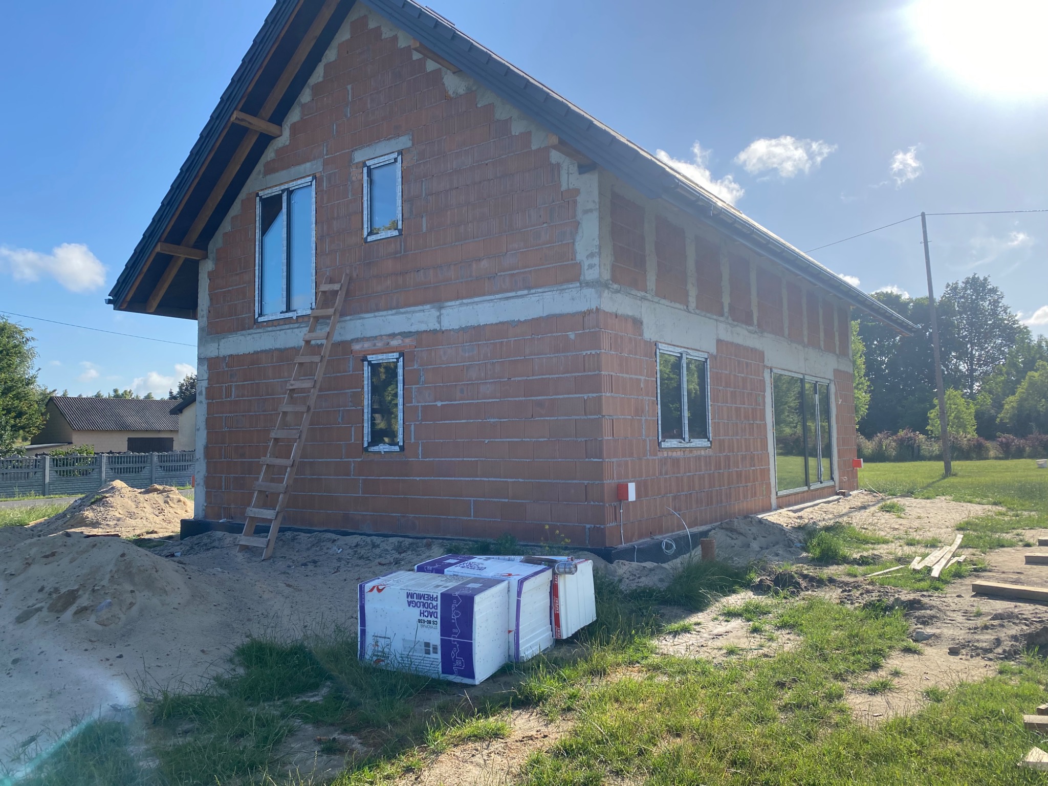 Surowy budynek z cegły z zamontowanymi oknami, drabina oparta o ścianę, materiały izolacyjne na trawie. Budowa domu jednorodzinnego w trakcie realizacji.