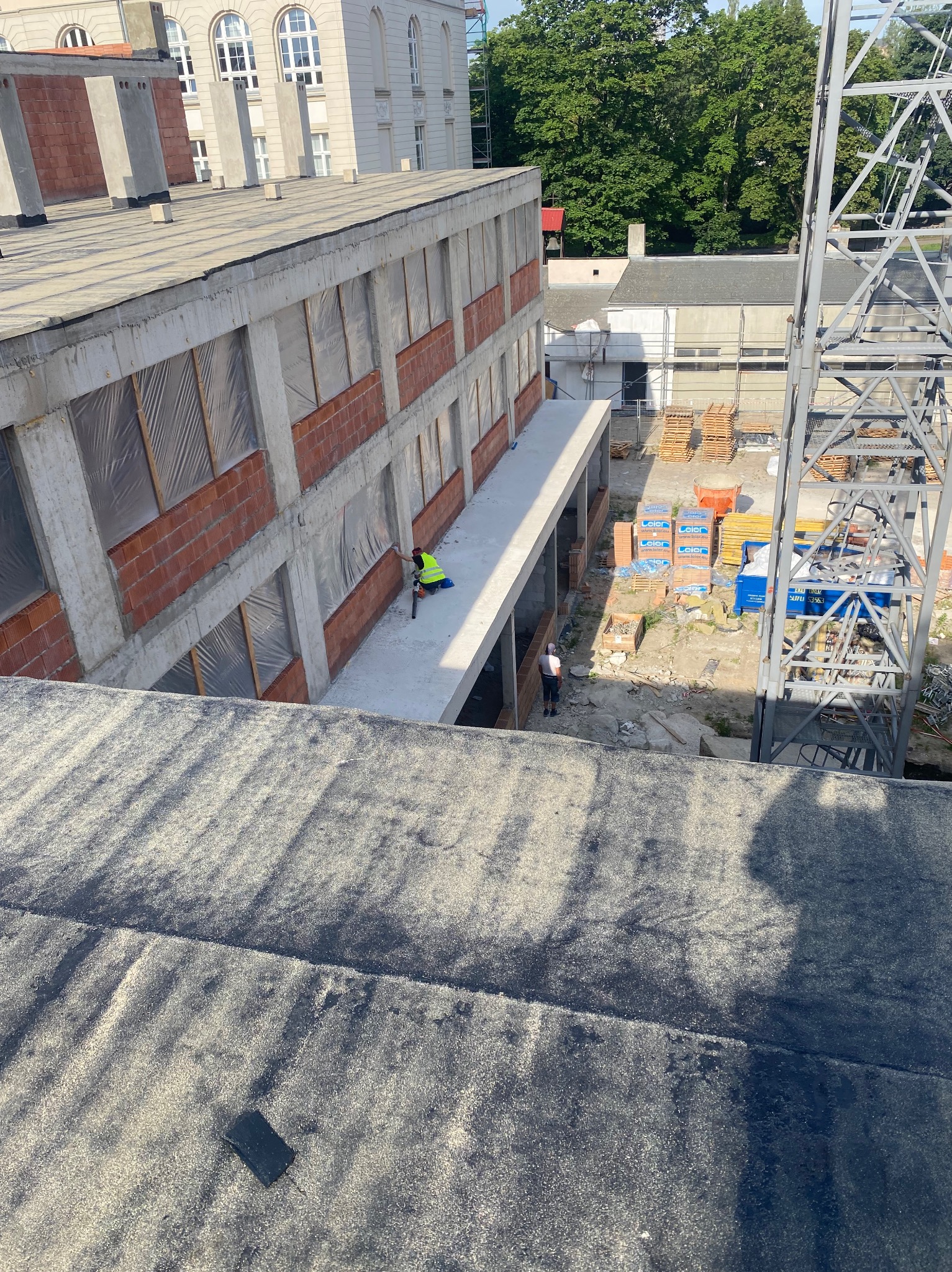 Wysoki kąt: Murarz w kamizelce, w trakcie prac murarskich na betonowym balkonie budynku w stanie surowym, z widoczną konstrukcją żurawia budowlanego.