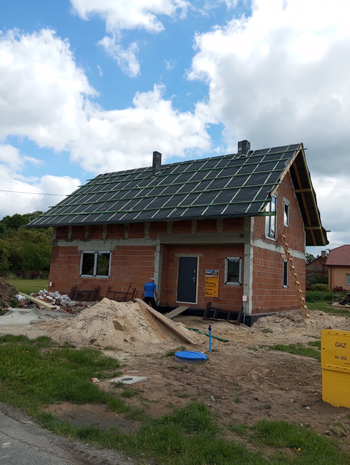 Dom w trakcie budowy: murowana konstrukcja, przygotowany dach z łatami, okna wstawione, teren wokół w piasku i gruzie, błękitne niebo z chmurami w tle.