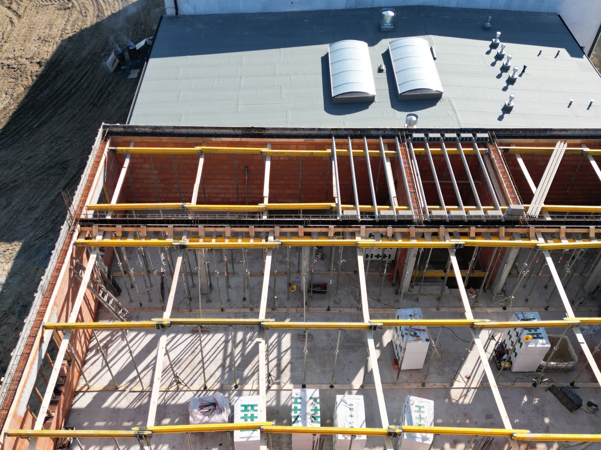 Budowa domu z cegły, widok z góry. Wnętrze z betonową podłogą, podparte żółtymi belkami i metalowymi podporami. Na dachu świetliki i kominy wentylacyjne.