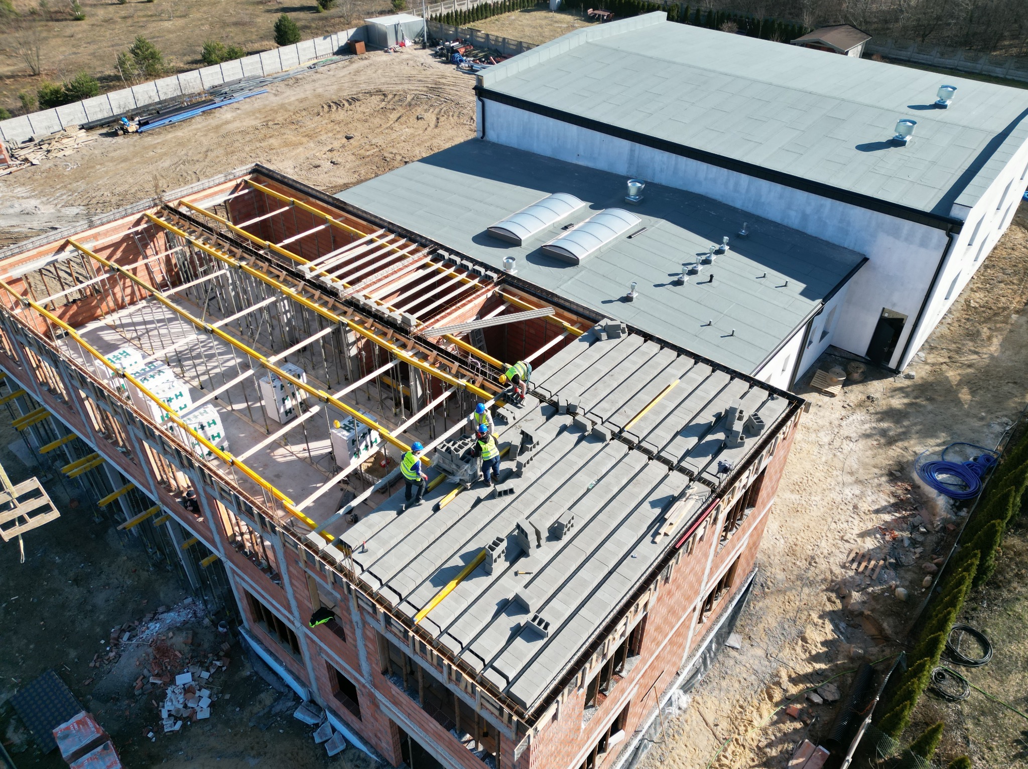 Budowa dwukondygnacyjnego budynku z cegły. Ekipa w kaskach układa prefabrykaty stropowe. Widok z góry na plac budowy i sąsiedni budynek z szarym dachem.