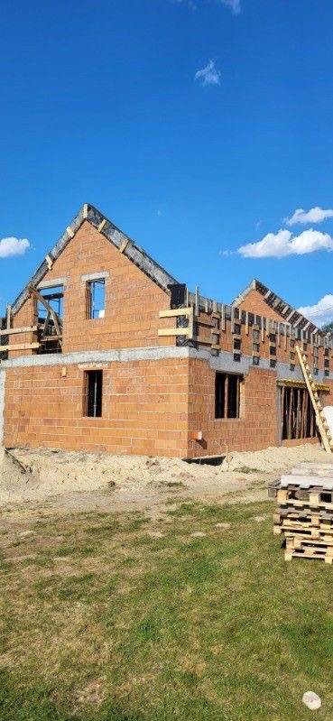 Surowy stan budynku w trakcie budowy, z ceglanymi ścianami i drewnianą konstrukcją dachu na tle błękitnego nieba z chmurami. Widoczne palety z materiałami budowlanymi.