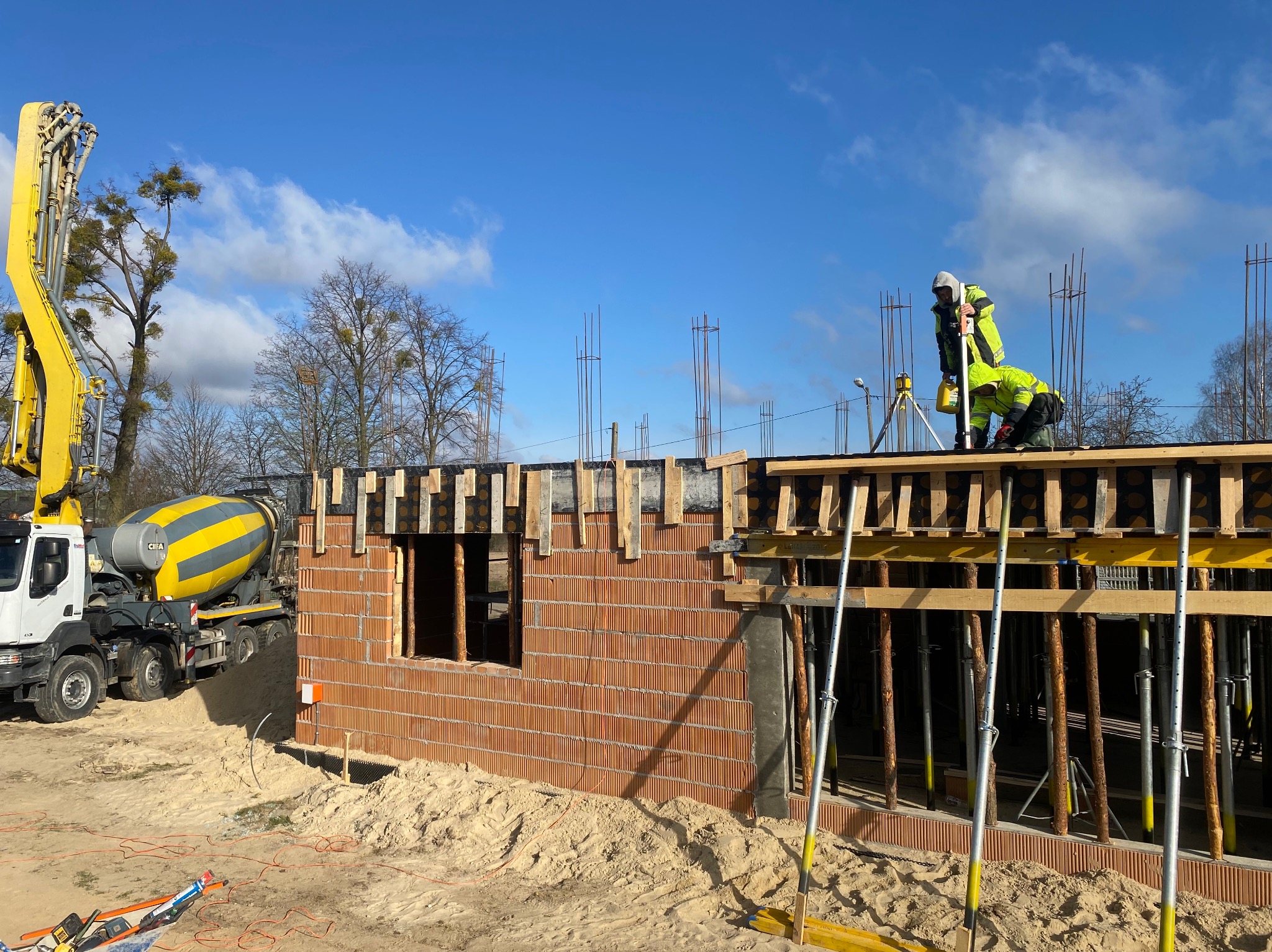 Budowa domu z betonu komórkowego: murarz w odblaskowej odzieży mierzy poziom, w tle pompa do betonu i błękitne niebo. Widoczne zbrojenia i drewniane szalunki.