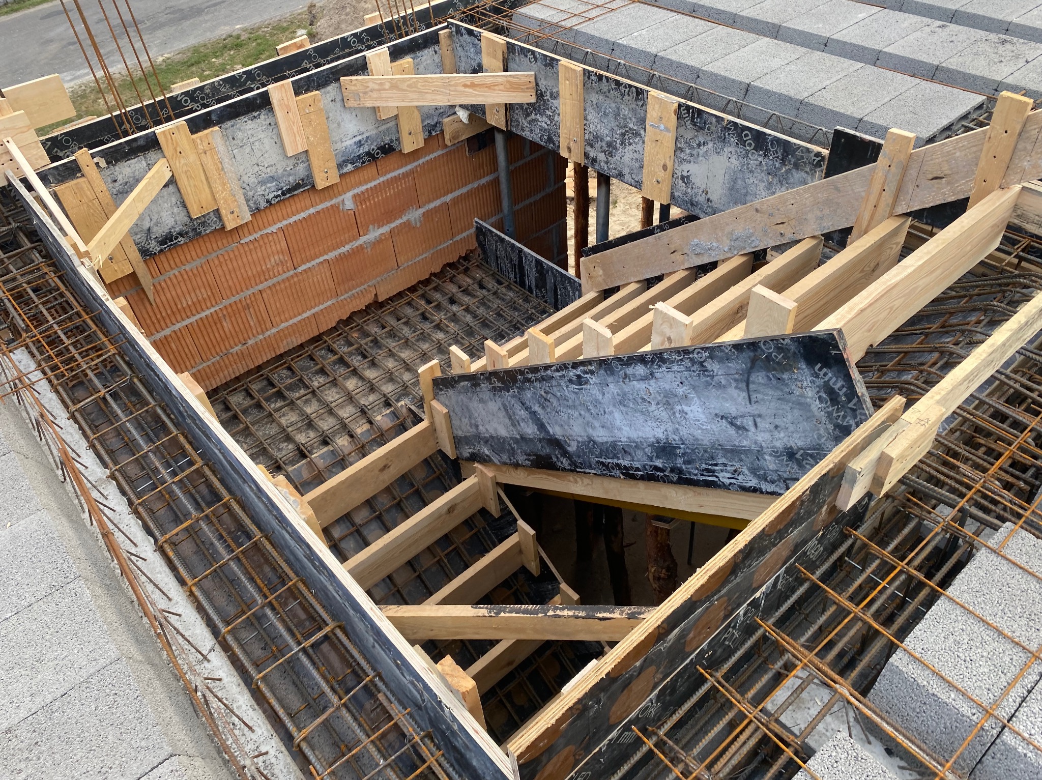 Zbrojenie stropu budynku w trakcie budowy, widok z góry. Konstrukcja z belek drewnianych, cegieł i betonu komórkowego. Elementy szalunku i siatka z prętów.