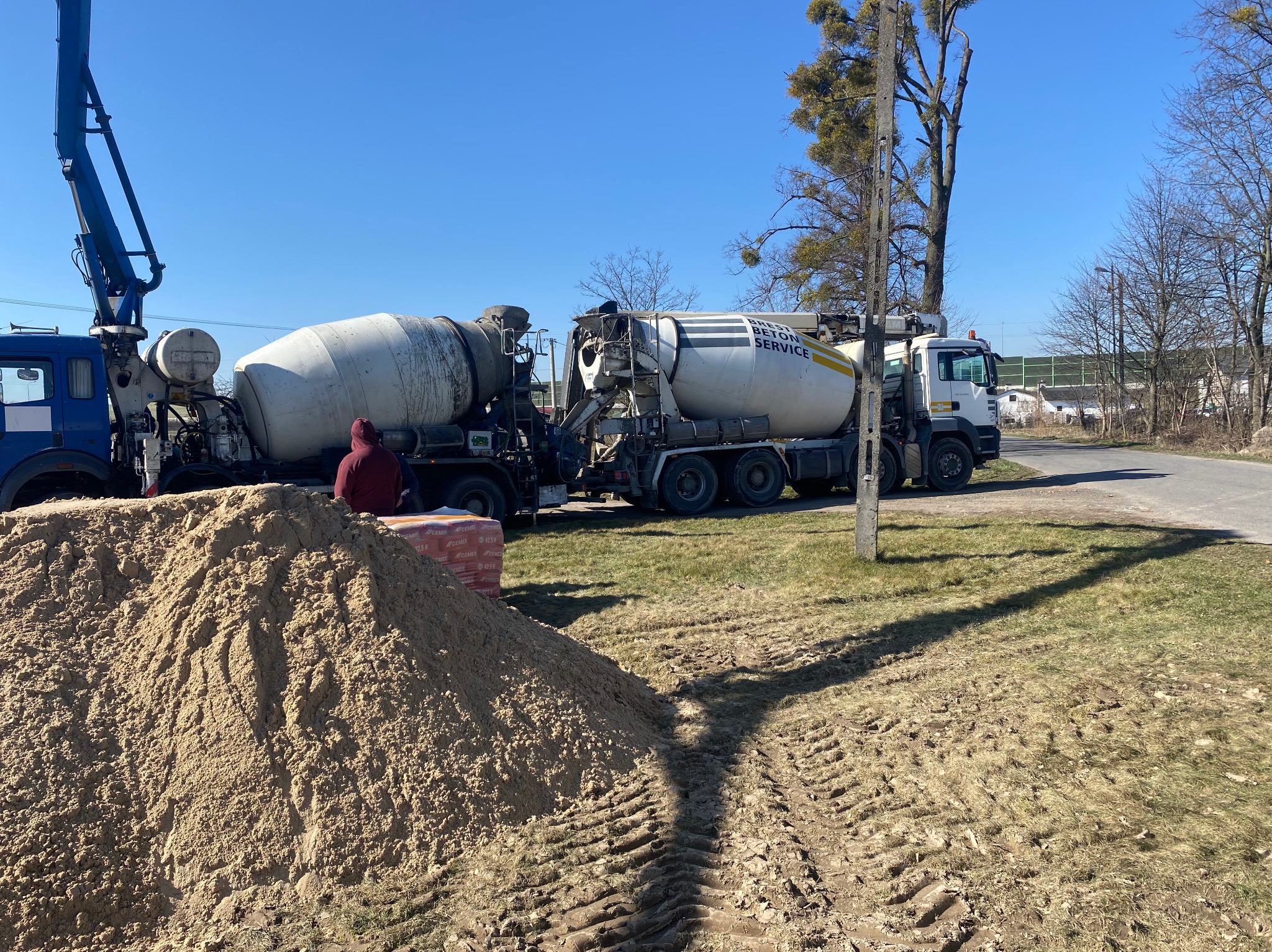 Dwa betonomieszarki obok sterty piasku i palety cegieł na trawie, w tle droga i drzewa pod błękitnym niebem. Widoczne ślady opon na trawie.
