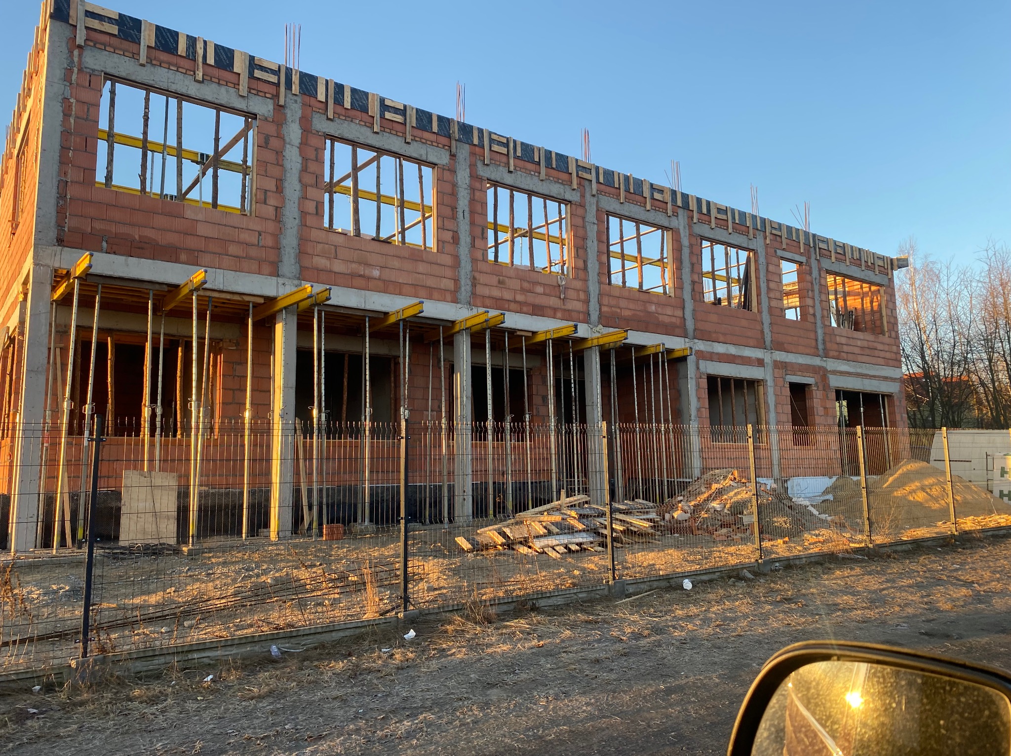 Dwukondygnacyjny budynek w stanie surowym otwartym z czerwonej cegły, widoczne podpory i szalunki okienne, ogrodzenie tymczasowe, wczesne stadium budowy.