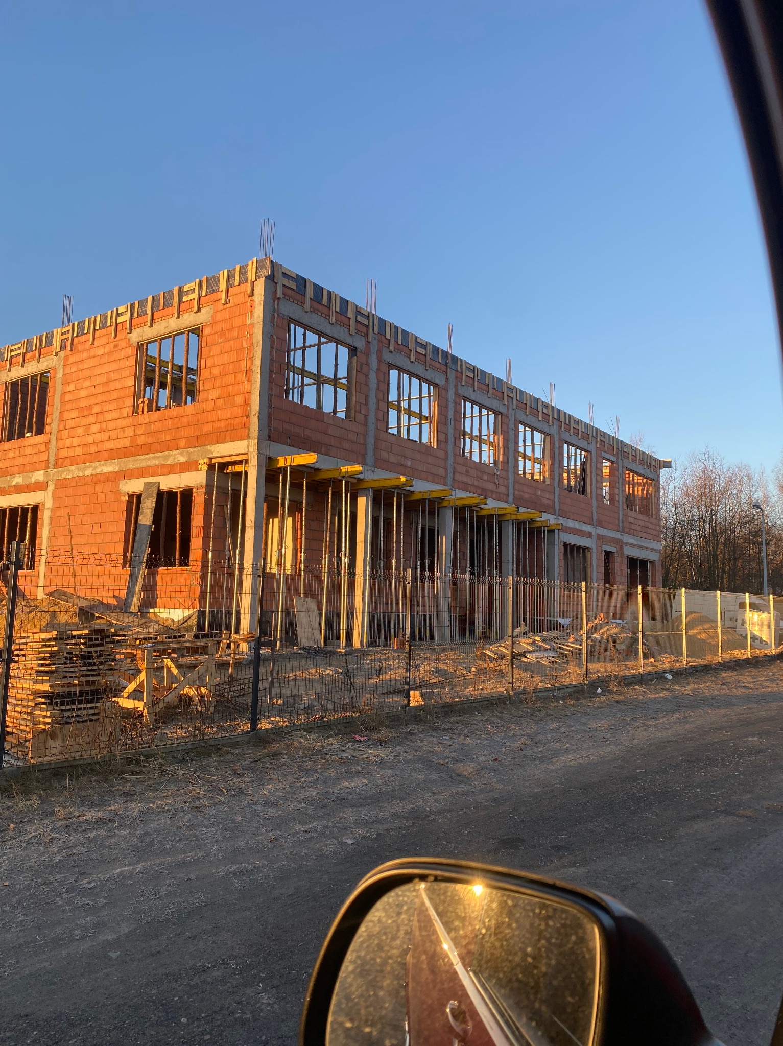 Wznoszenie dwukondygnacyjnego budynku z czerwonej cegły, widoczne szalunki i podpory. Częściowo ogrodzony teren budowy w słoneczny dzień.