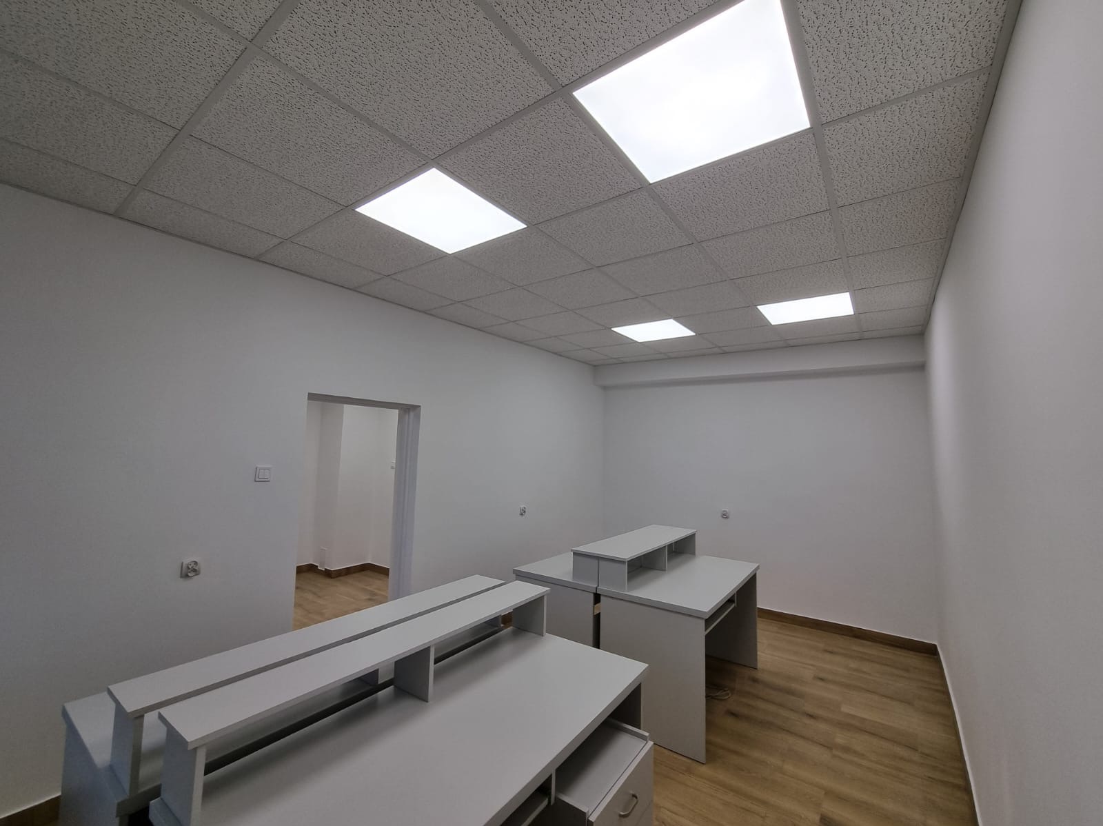Wnętrze biura po malowaniu w Łodzi: białe ściany, podwieszany sufit z oświetleniem, jasne biurka, drewnopodobna podłoga. Widok na odnowione pomieszczenie.