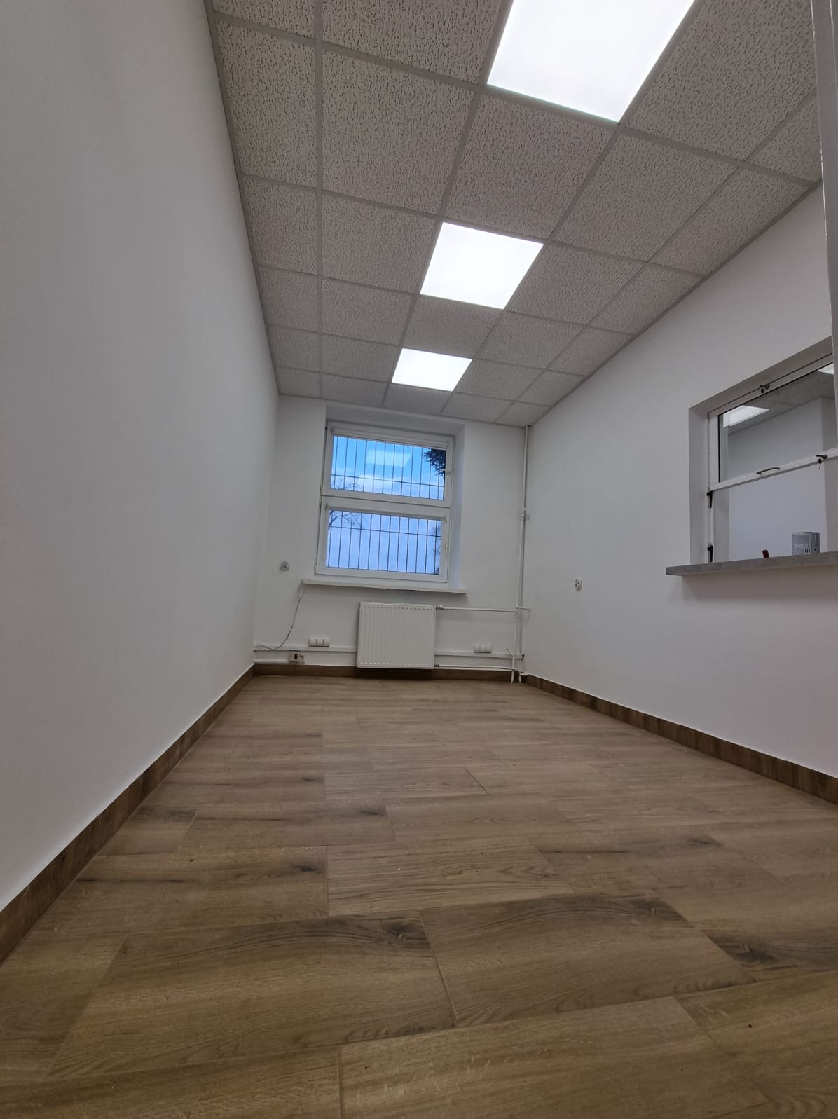 Wnętrze biura po remoncie: świeżo pomalowane ściany na biało, drewnopodobna podłoga, okno z kratą i grzejnikiem, sufit kasetonowy z oświetleniem.