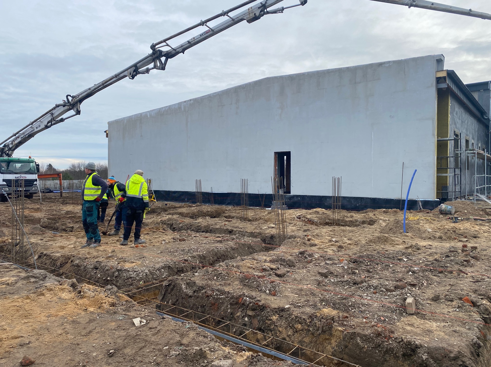 Budowa hali: fundamenty zbrojone stalą, pracownicy w odblaskowych kamizelkach, betonowa pompa w tle. Etap wylewania fundamentów pod budynek.