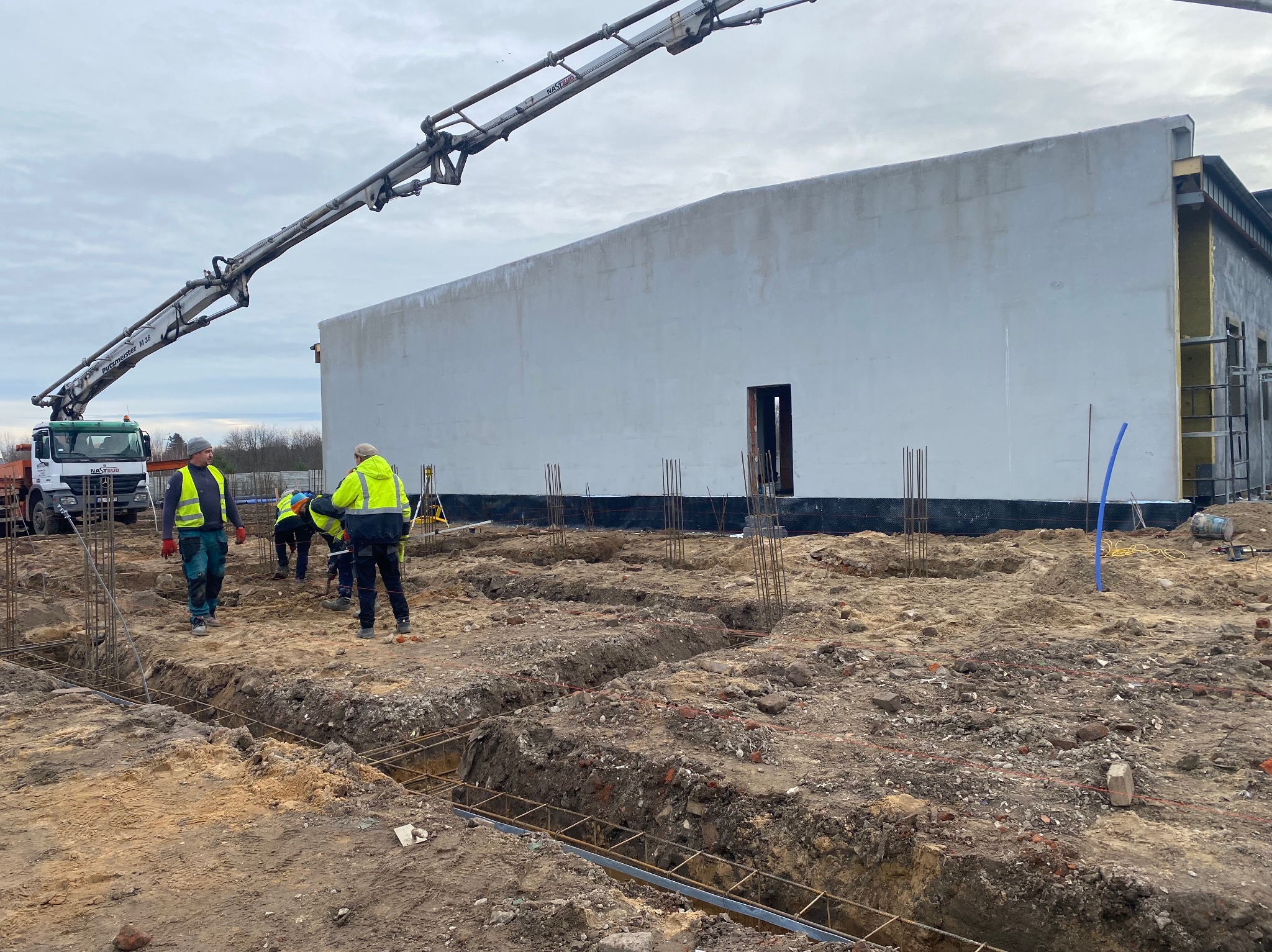Budowa fundamentów budynku: ekipa w odblaskowych kamizelkach, betonowa pompa, wykopane rowy zbrojeniowe, w tle surowa ściana budynku. Prace ziemne w toku.