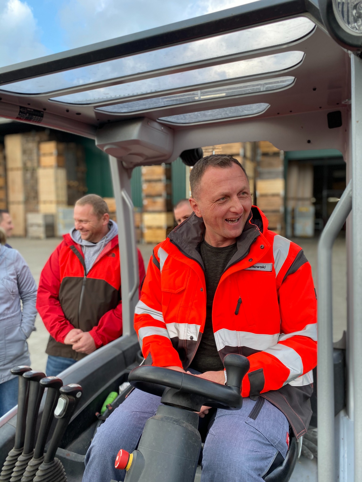 Uśmiechnięty operator wózka widłowego w pomarańczowej kurtce odblaskowej, siedzący za sterami w kabinie, w tle grupa obserwujących osób i palety z drewnem.