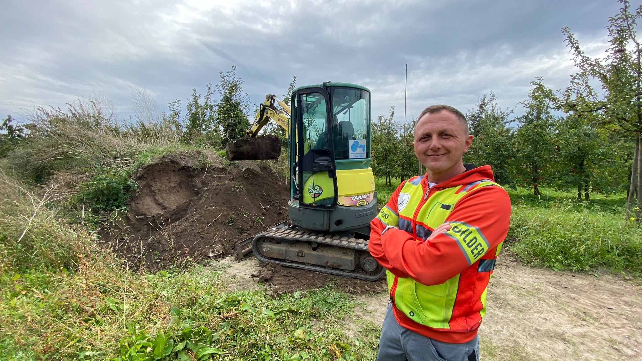 Operator w kamizelce odblaskowej uśmiecha się na tle pracującej mini-koparki Yanmar w Guderhandviertel. Koparka nabiera ziemię z wykopu na terenie sadu.