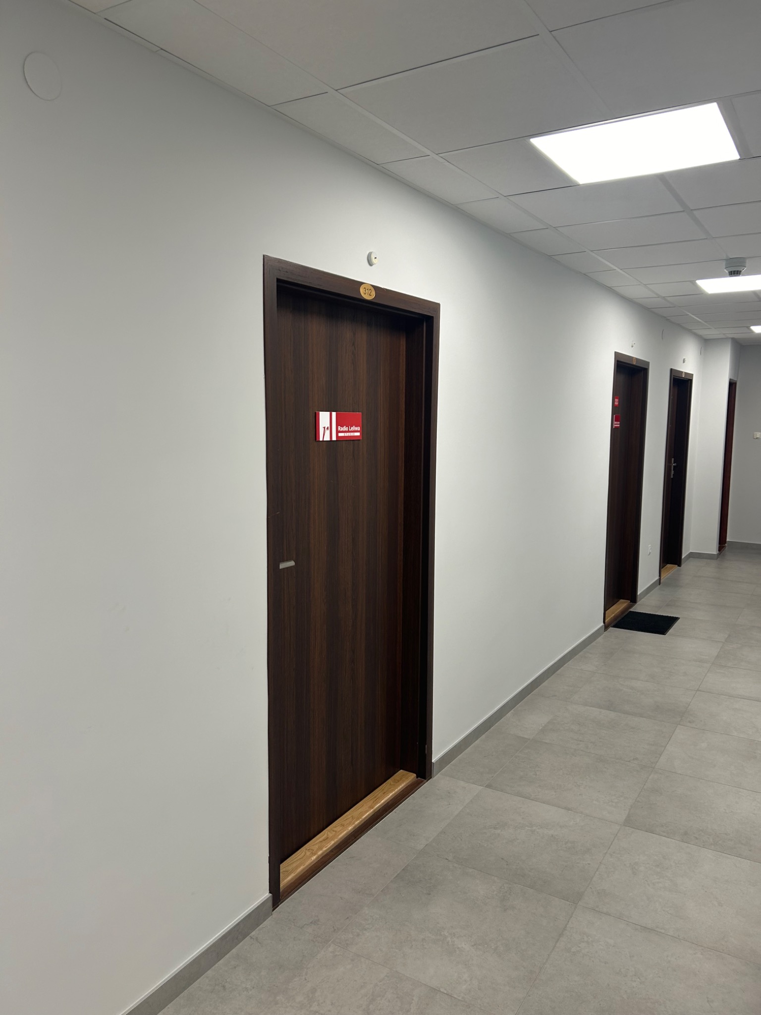 Korytarz biurowy z białymi ścianami, szarą podłogą z płytek i ciemnobrązowymi drzwiami z numerami pokoi i logo firmy.
