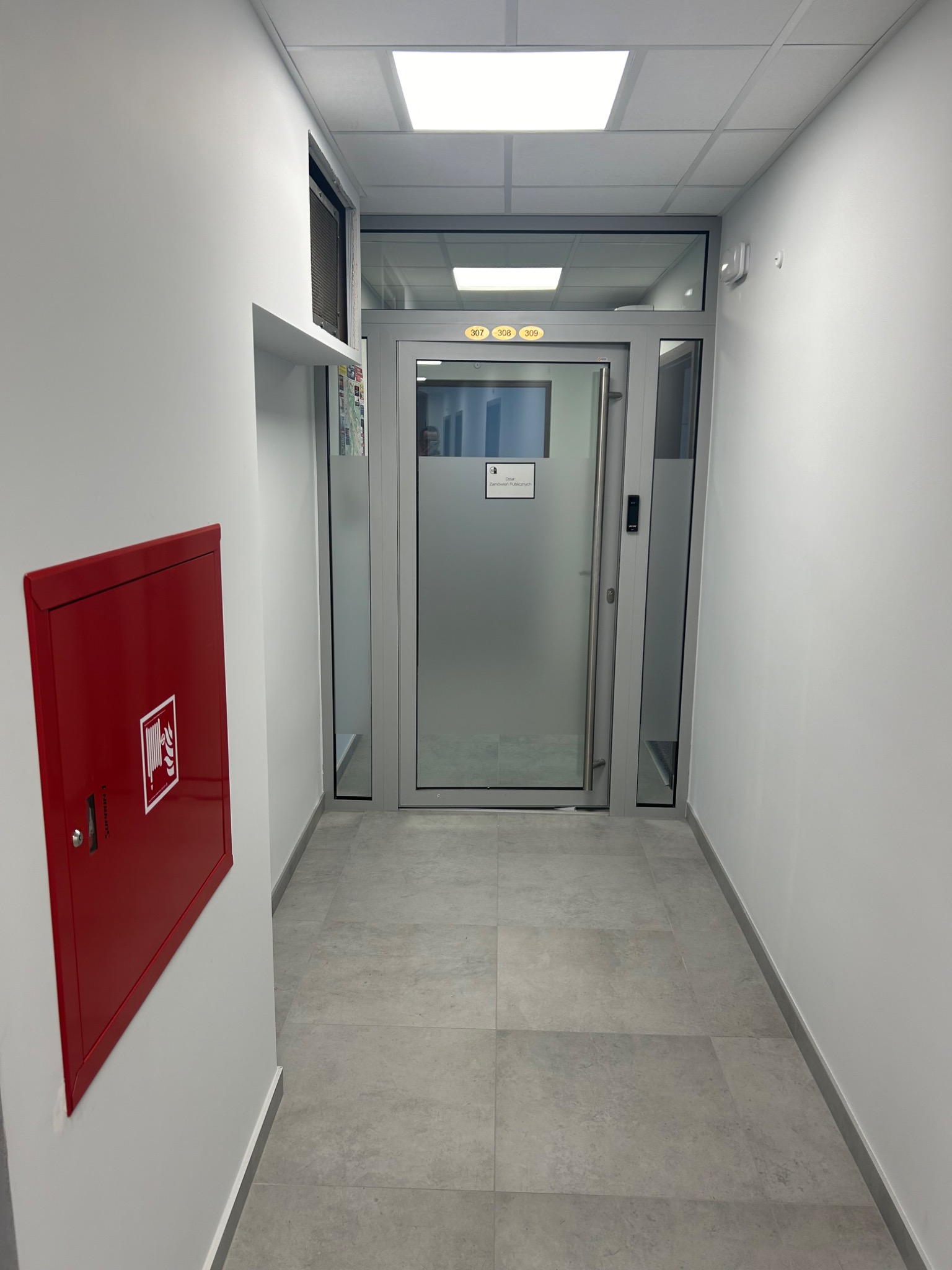 Korytarz biurowy w Stalowej Woli z pomalowanymi na biało ścianami, szarą podłogą z płytek, stalowymi drzwiami z numerami pokoi 307, 308, 309 i czerwoną szafką hydrantową.