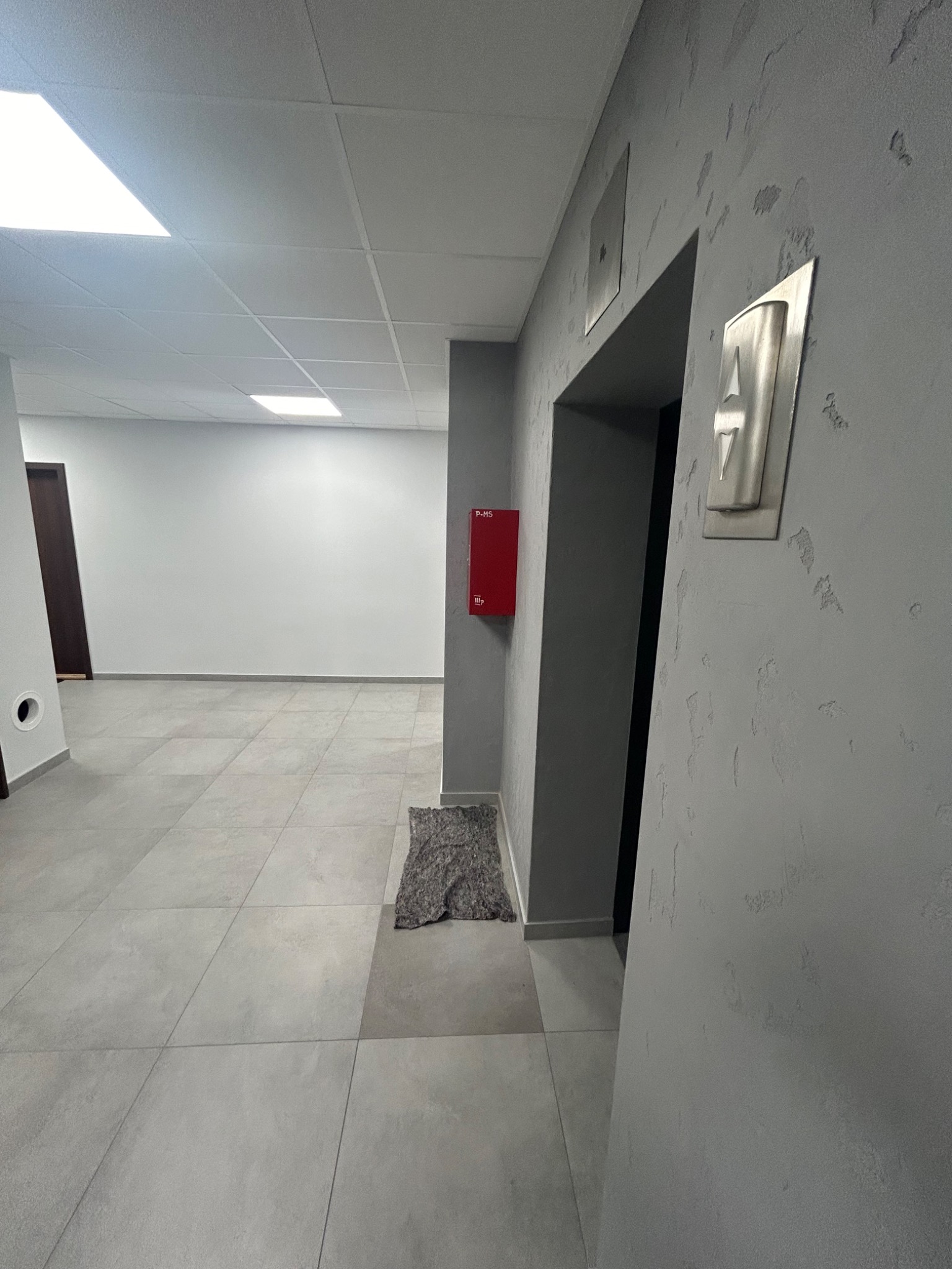 Szary korytarz biurowy z dekoracyjną fakturą na ścianach, widoczne płytki podłogowe, oświetlenie sufitowe, drzwi i czerwona skrzynka hydrantowa.