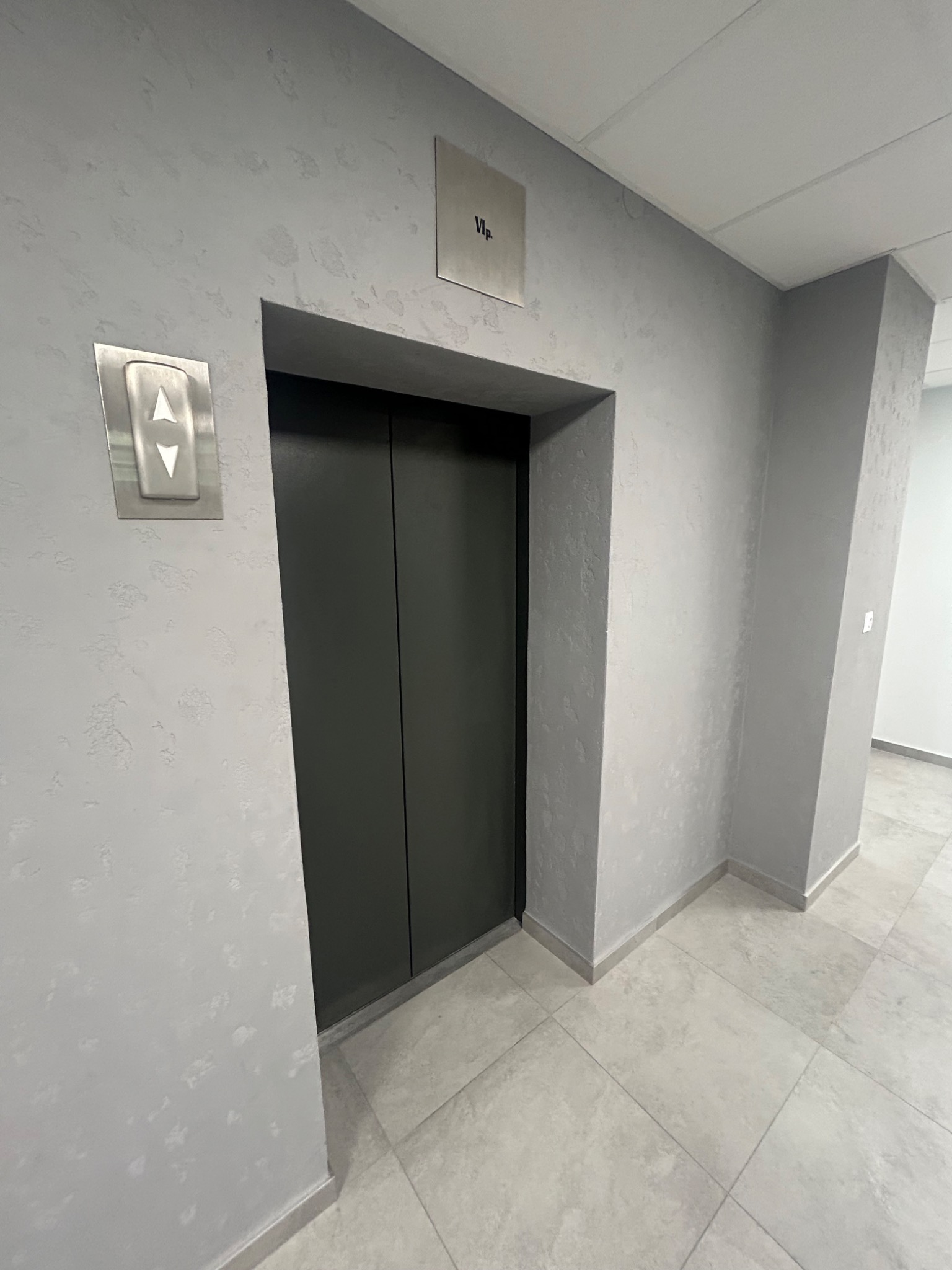 Szare ściany z dekoracyjną fakturą w korytarzu biurowym, obok zamkniętych drzwi windy z metalowym panelem sterowania.