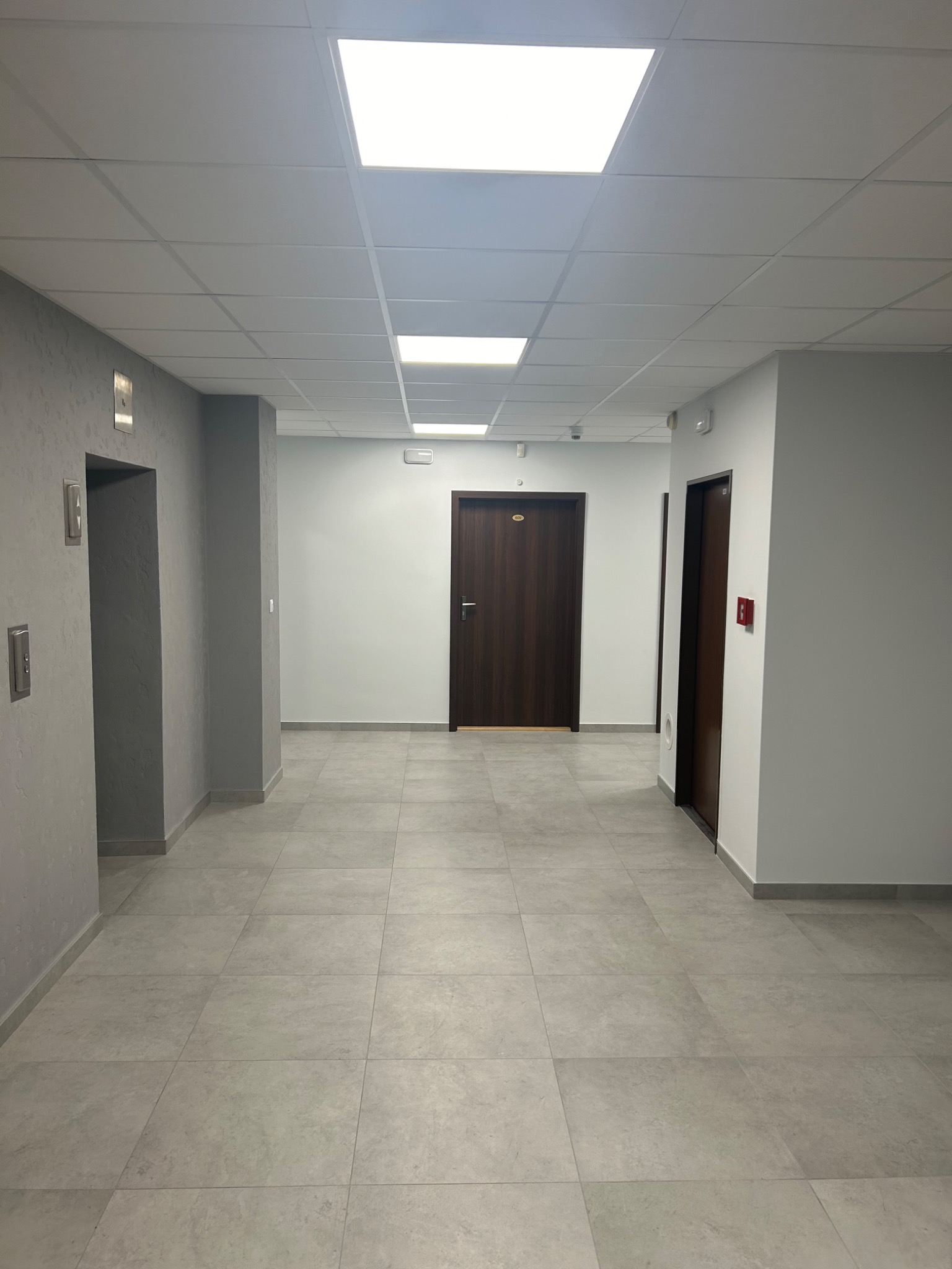 Korytarz biurowy z szarą podłogą z płytek, białymi ścianami, drzwiami z numerami pokoi i podwieszanym sufitem z panelami oświetleniowymi.