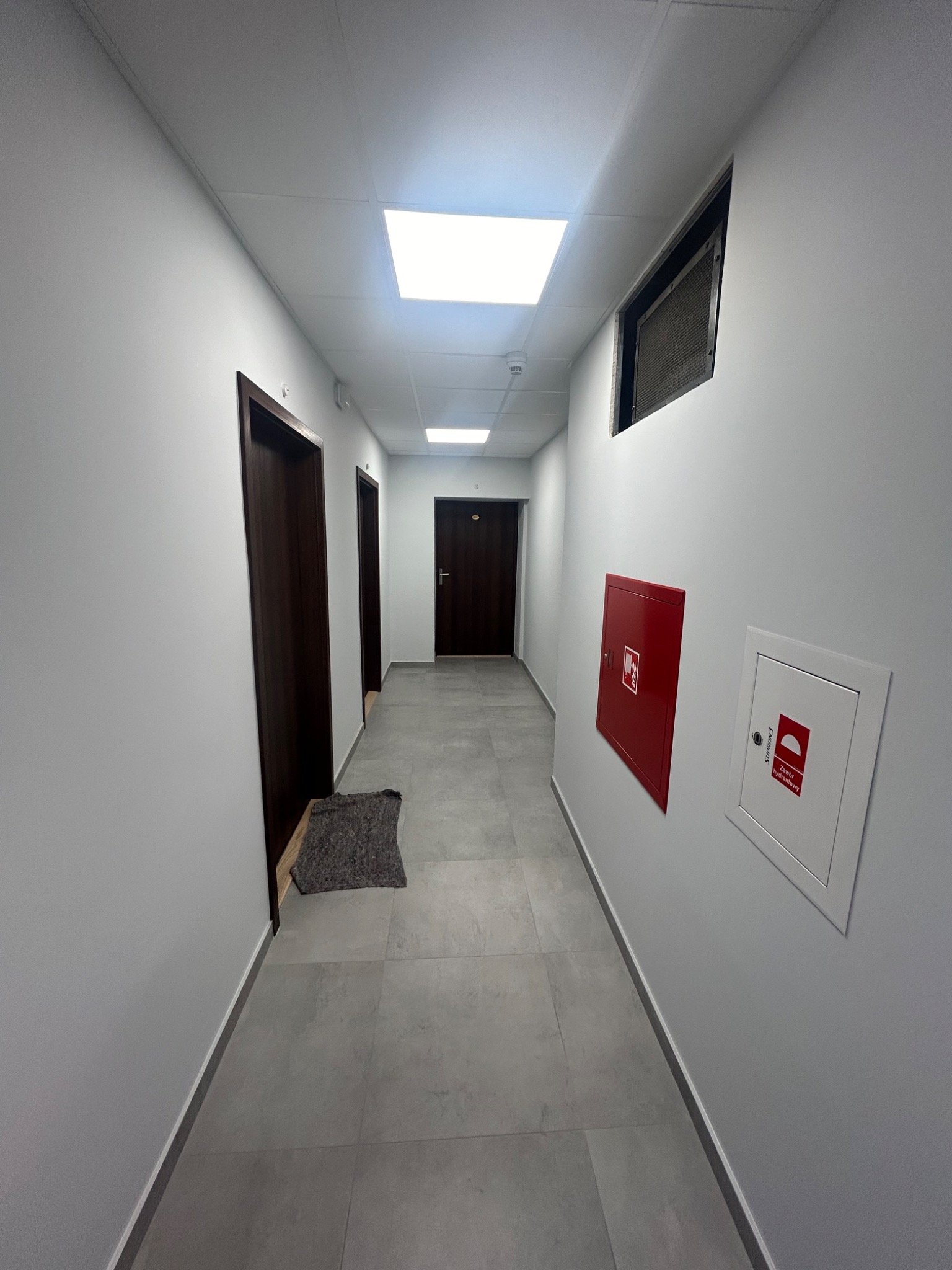 Korytarz biurowy z szarymi płytkami podłogowymi, białymi ścianami, brązowymi drzwiami, czerwonym hydrantem i kratką wentylacyjną pod sufitem.