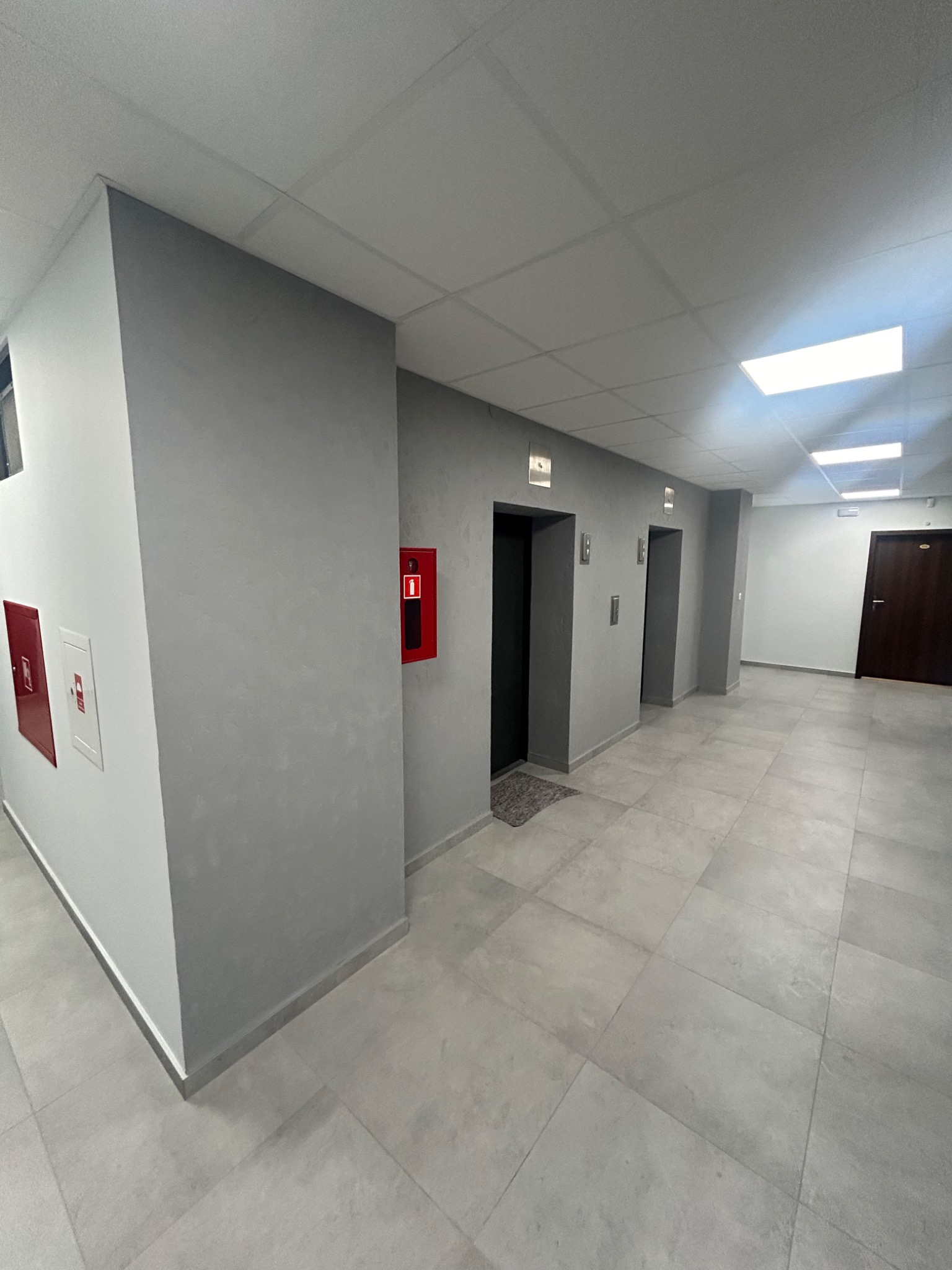 Szary korytarz biurowy z trzema windami, czerwonymi szafkami hydrantowymi i ciemnymi drzwiami na końcu, oświetlony panelami LED w suficie.