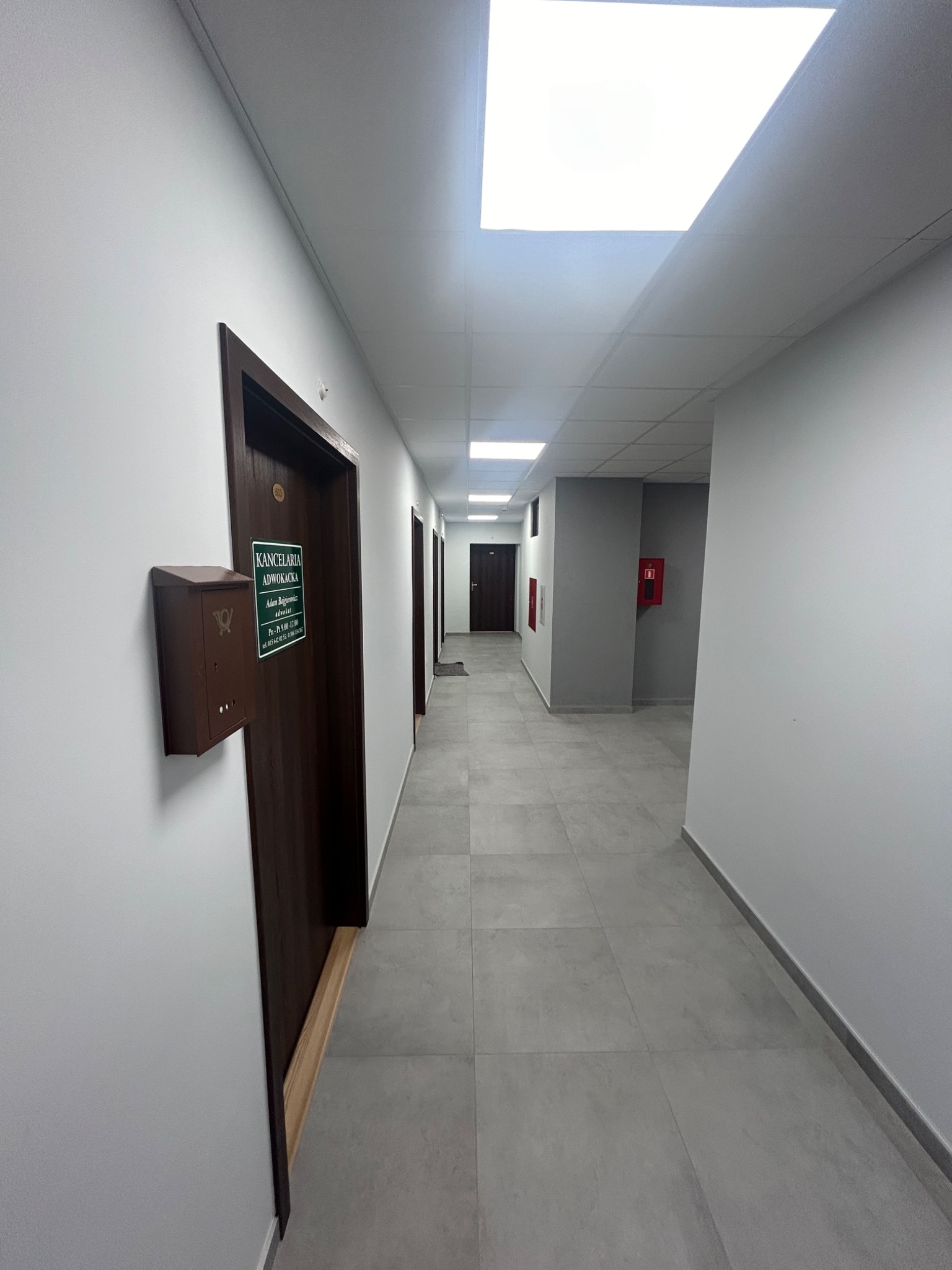 Długi korytarz biurowy z szarymi płytkami podłogowymi, białymi ścianami i ciemnymi drzwiami z tabliczkami informacyjnymi. W tle widoczny czerwony hydrant.
