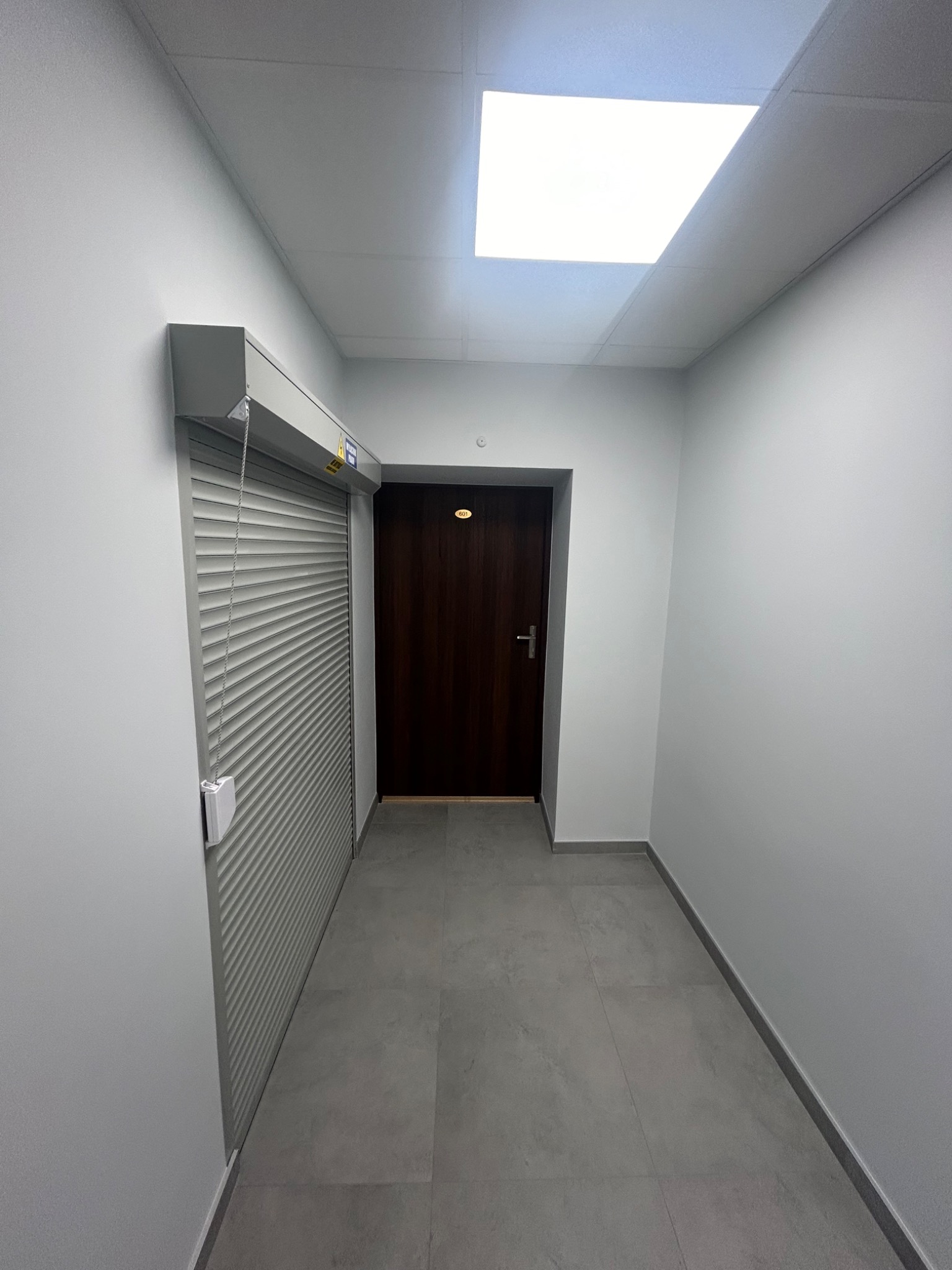 Korytarz biurowy z szarą podłogą, białe ściany i ciemne drzwi z numerem pokoju, zamknięta roleta antywłamaniowa przy ścianie.