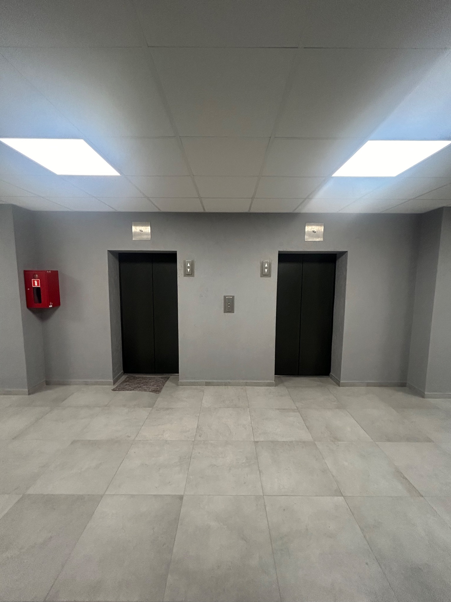 Szare wnętrze korytarza z dwoma windami, oświetleniem sufitowym i czerwoną gaśnicą na ścianie.