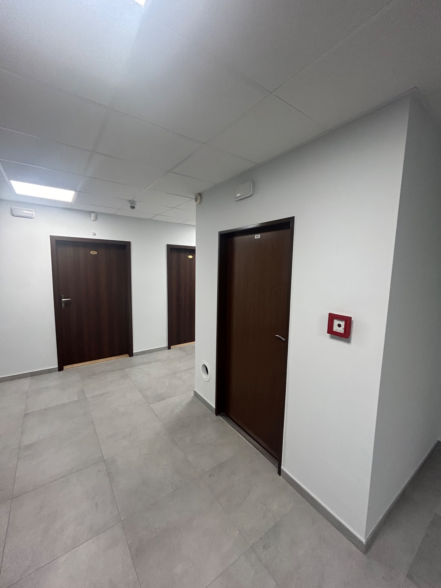 Korytarz biurowy z szarymi płytkami podłogowymi, białymi ścianami, brązowymi drzwiami z numerami i systemem przeciwpożarowym.