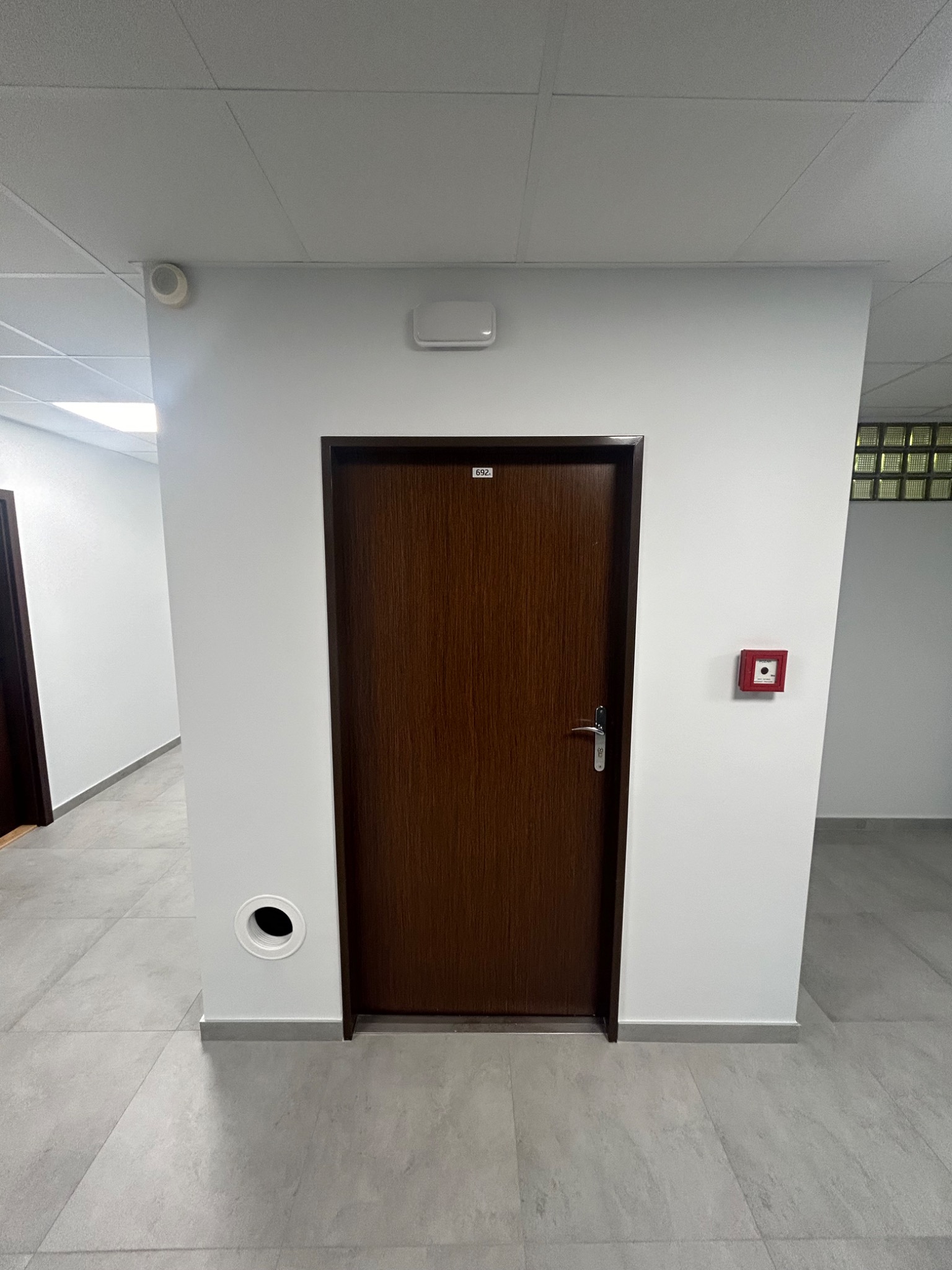 Wnętrze biura z ciemnobrązowymi drzwiami o numerze 692, białą ścianą z czujnikiem dymu i czerwoną skrzynką alarmową, szarą podłogą z płytek i białym sufitem podwieszanym.