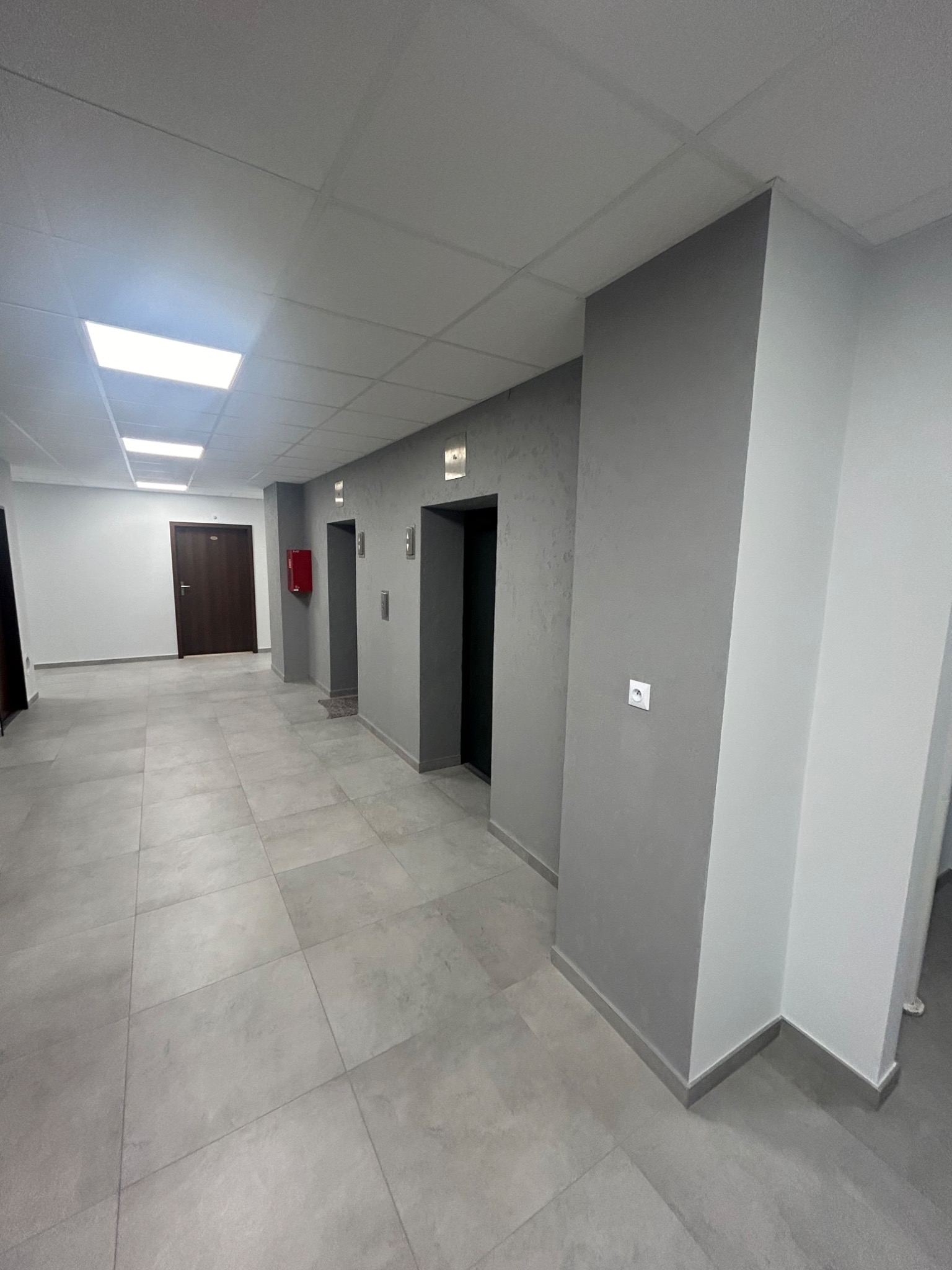 Wnętrze korytarza biurowego z szarymi ścianami, jasnymi płytkami podłogowymi, sufitami podwieszanymi z panelami LED i windami po prawej stronie.