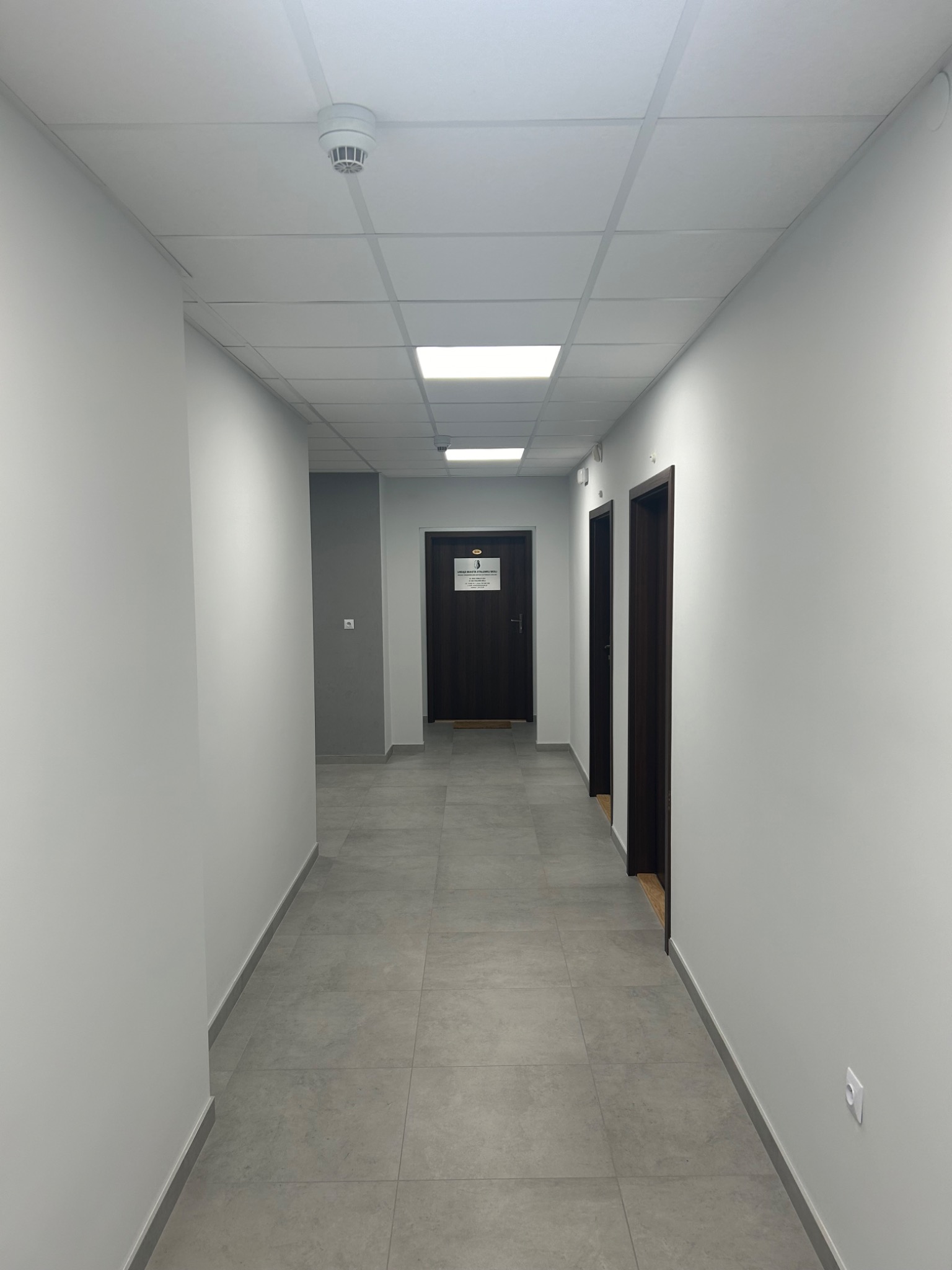 Długi korytarz biurowy z szarymi płytkami podłogowymi, białymi ścianami i ciemnymi drzwiami, oświetlony panelami LED w suficie kasetonowym.