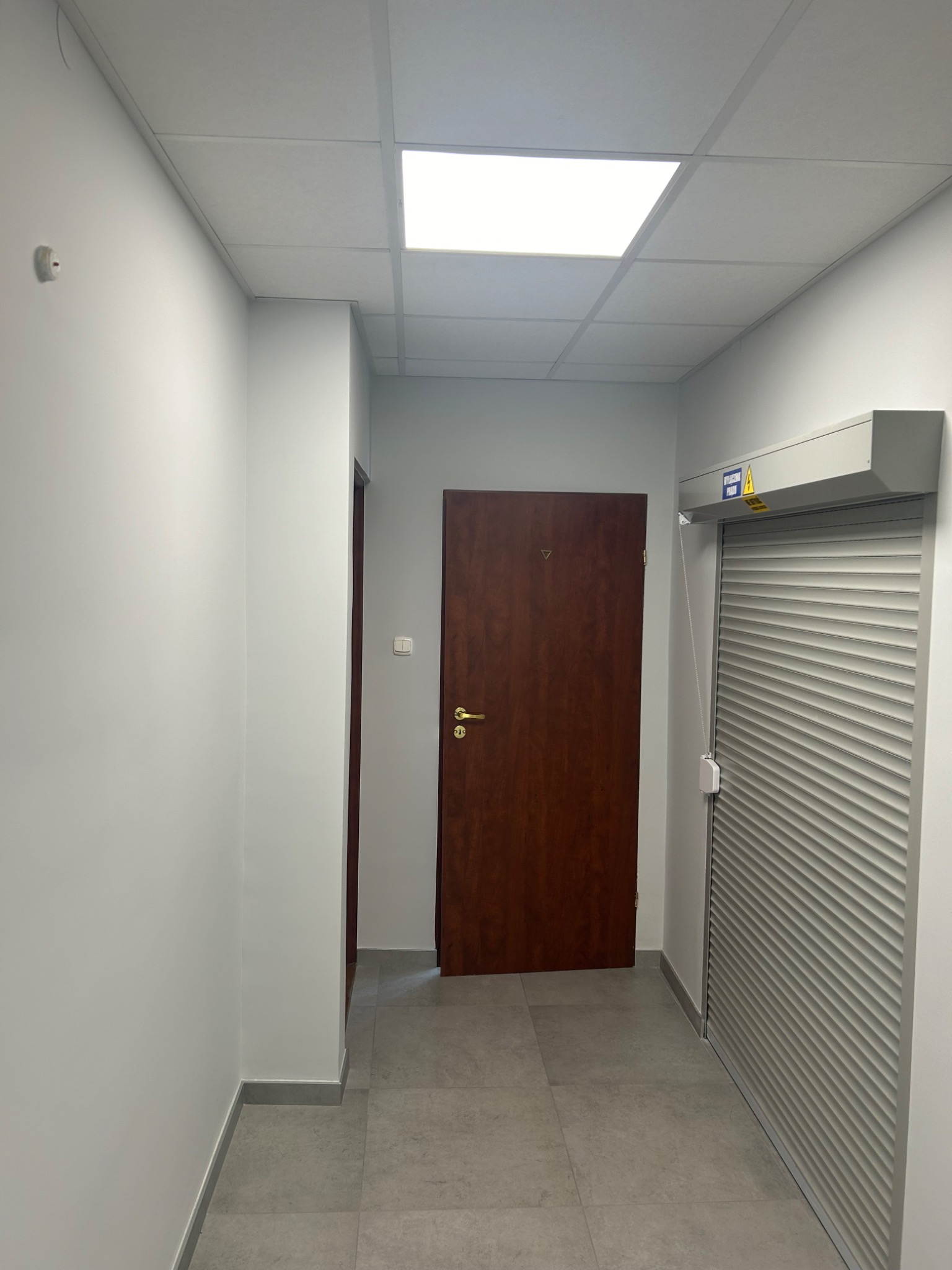 Korytarz biurowy z szarą podłogą, białymi ścianami, drewnianymi drzwiami i metalową roletą zabezpieczającą, oświetlony panelami sufitowymi.