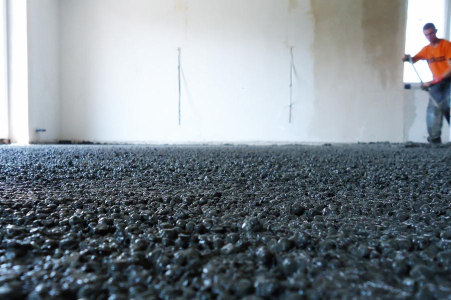 Szara, mokra wylewka na podłodze w trakcie wykonywania, widoczny pracownik zacierający powierzchnię w jasnym pomieszczeniu z surowymi ścianami.