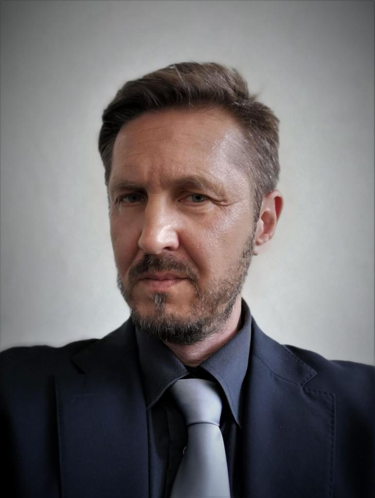 Portret mężczyzny w ciemnym garniturze i szarym krawacie, z lekkim zarostem, na jasnoszarym tle.