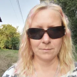 Portret kobiety w okularach przeciwsłonecznych na tle zieleni. Blond włosy z różowymi pasemkami, srebrny łańcuszek. Ujęcie z dołu, widoczne drzewa i niebo.