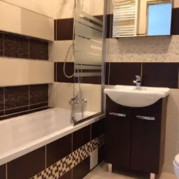 Wykończona łazienka z brązowo-beżowymi płytkami, wanną z szklanym parawanem, szafką z umywalką i grzejnikiem drabinkowym.