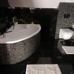 Luksusowa łazienka z czarnymi marmurowymi ścianami i podłogą, mozaikową obudową wanny, wiszącą toaletą i dekoracyjnym oświetleniem.